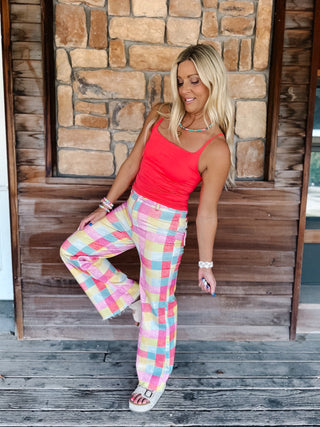 A woman posing on a rustic wooden porch with a stone wall background, wearing a bright coral red tank top and pastel pink, yellow, and blue plaid wide-leg pants.