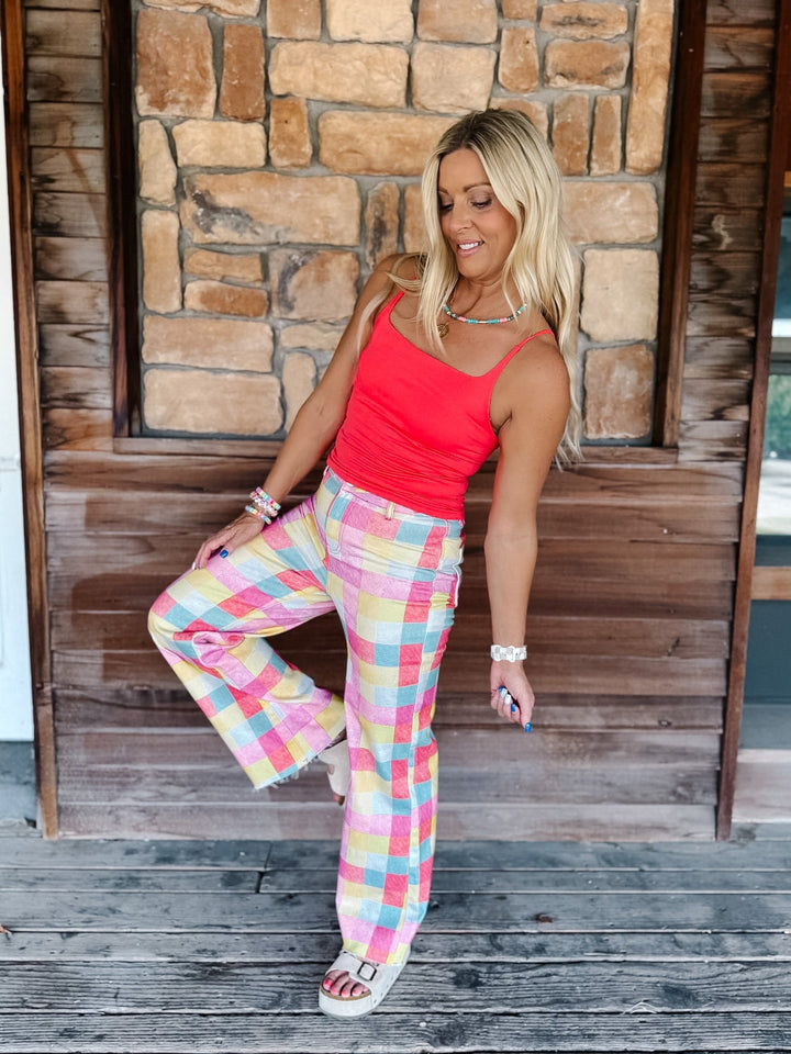 A woman posing on a rustic wooden porch with a stone wall background, wearing a bright coral red tank top and pastel pink, yellow, and blue plaid wide-leg pants.