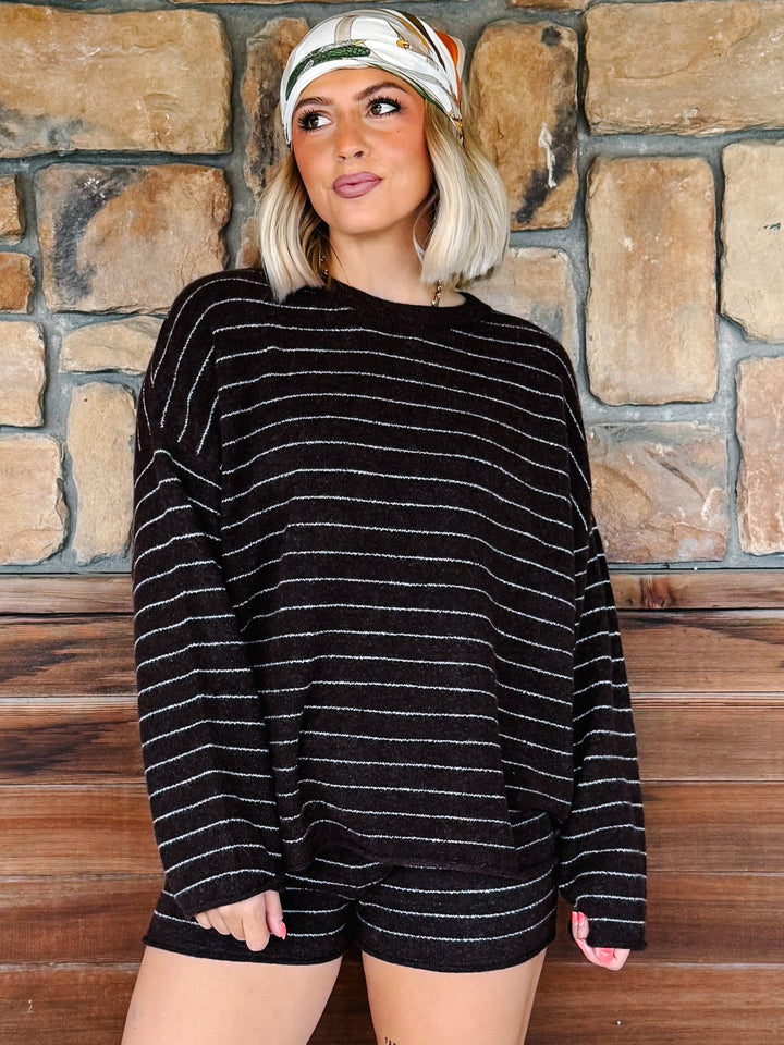 A woman wearing a brown knit long-sleeve top with thin white horizontal stripes and matching brown shorts, standing in front of a rustic stone wall and wooden bench.