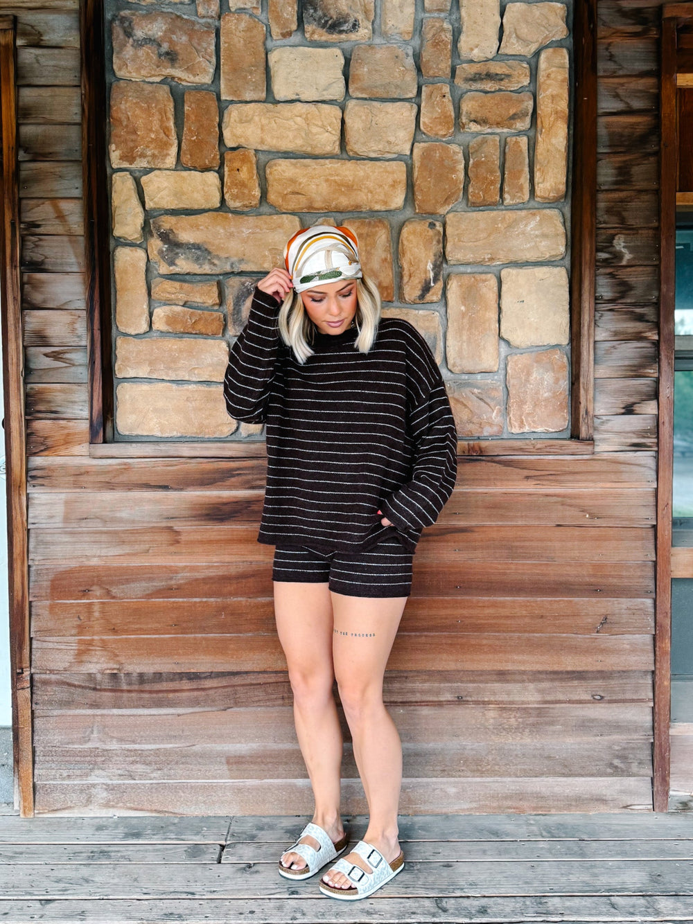 A woman stands on a wooden porch in front of a stone wall, wearing a matching brown and white striped long-sleeve top and shorts set, a striped headband, and light blue slide sandals.