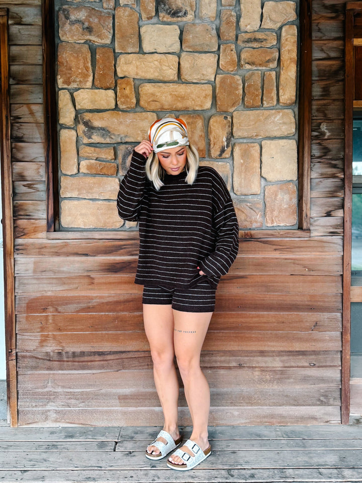 A woman stands on a wooden porch in front of a stone wall, wearing a matching brown and white striped long-sleeve top and shorts set, a striped headband, and light blue slide sandals.