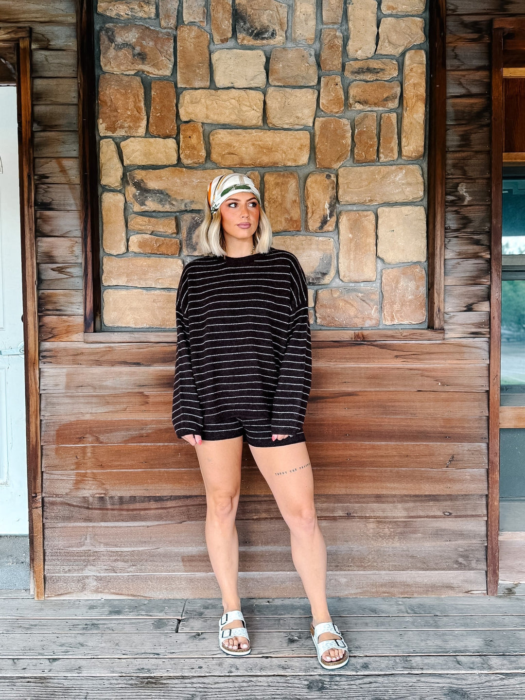 A woman stands on a rustic wooden porch in front of a stone wall, wearing a brown and white striped long-sleeve crop top and matching shorts, a patterned headband, and white slide sandals.