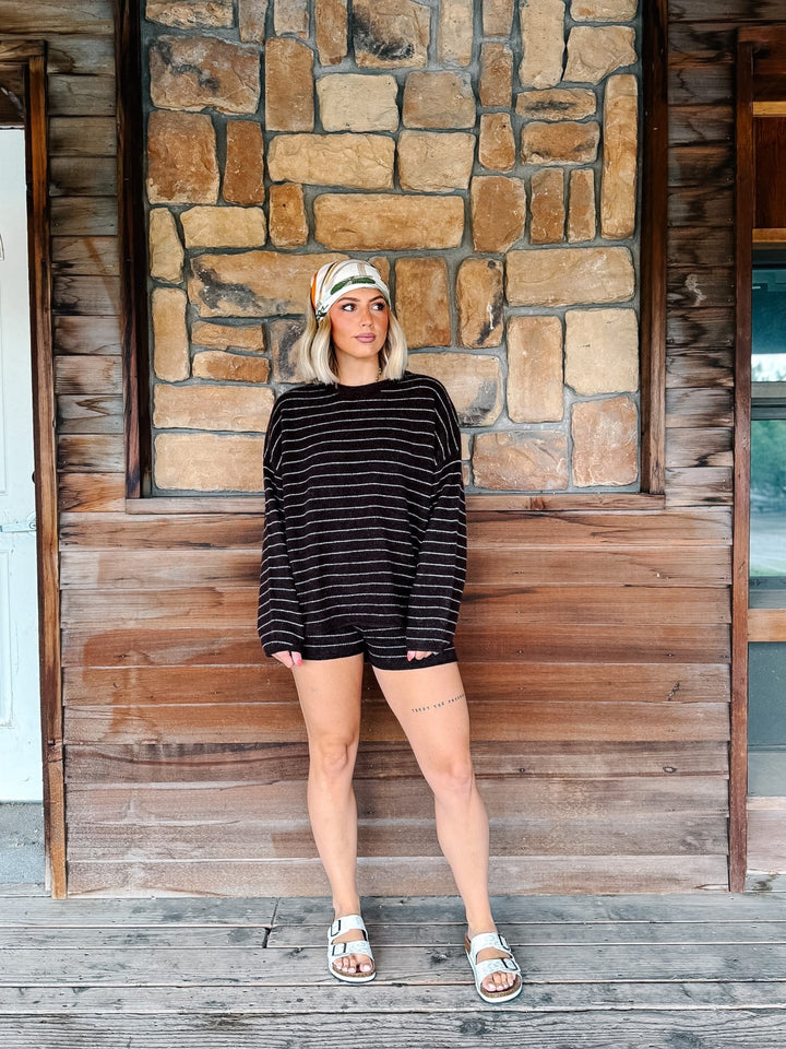 A woman stands on a rustic wooden porch in front of a stone wall, wearing a brown and white striped long-sleeve crop top and matching shorts, a patterned headband, and white slide sandals.