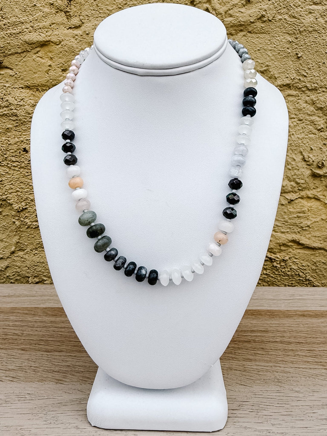 A beaded necklace displayed on a white jewelry bust, featuring alternating strands of polished black, white, pale pink, and gray-green beads, set against a textured tan wall and light wood surface.