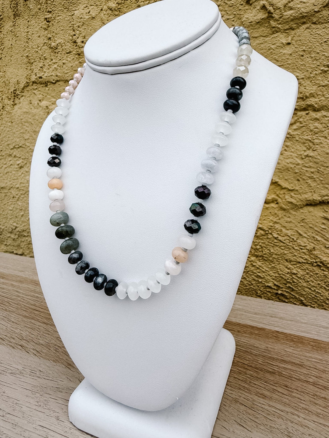 A multi-strand beaded necklace displayed on a white mannequin bust, featuring black, white, gray, and peach-colored round beads, set against a textured yellow wall and light wood surface.