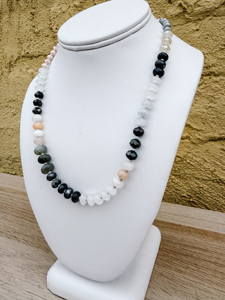 A multi-strand beaded necklace displayed on a white mannequin bust, featuring black, white, gray, and peach-colored round beads, set against a textured yellow wall and light wood surface.