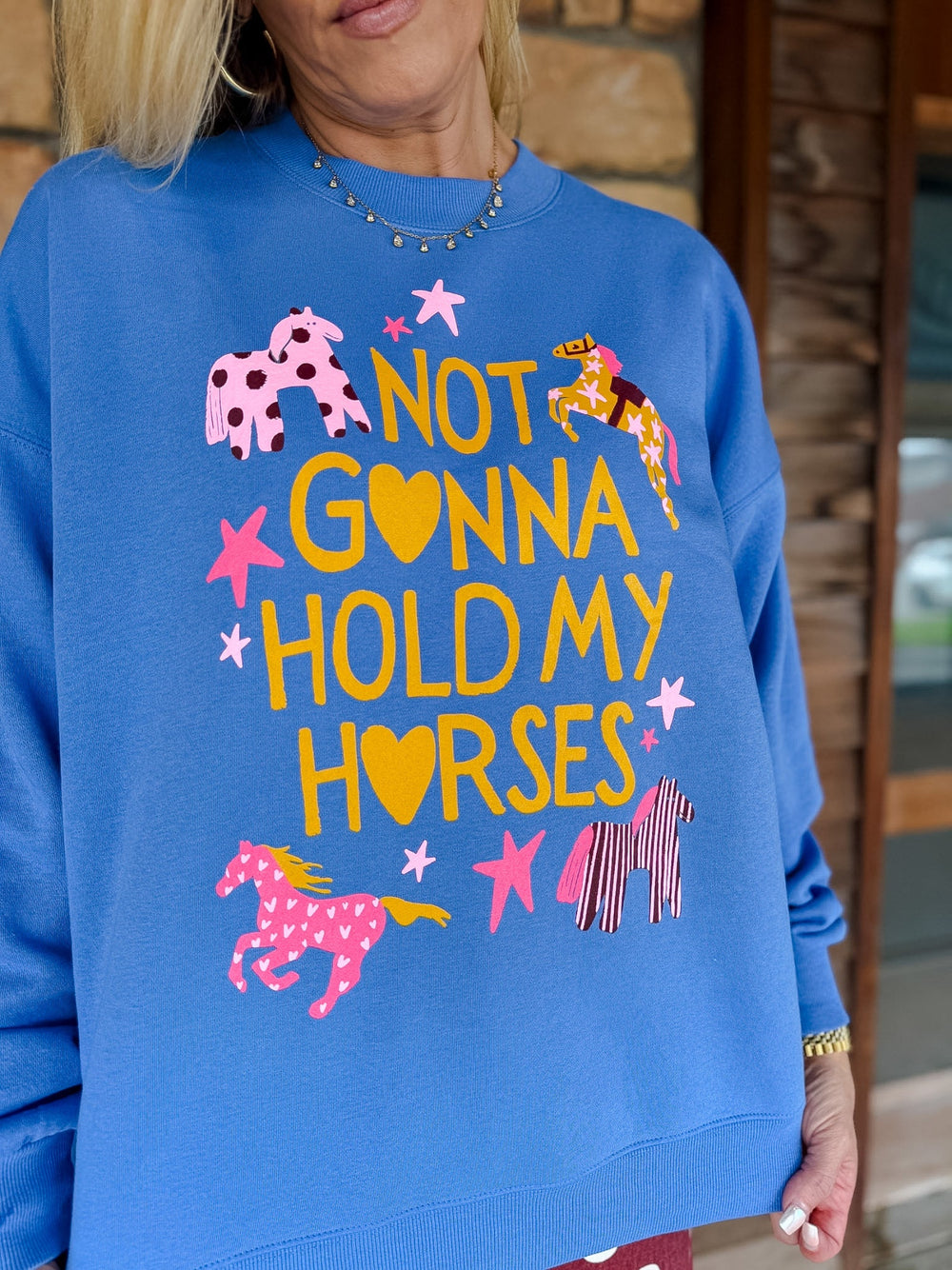 A woman wearing a bright blue crewneck sweatshirt with a colorful graphic that reads 'NOT GONNA HOLD MY HORSES' in yellow letters, surrounded by pink and yellow horse illustrations and star shapes.
