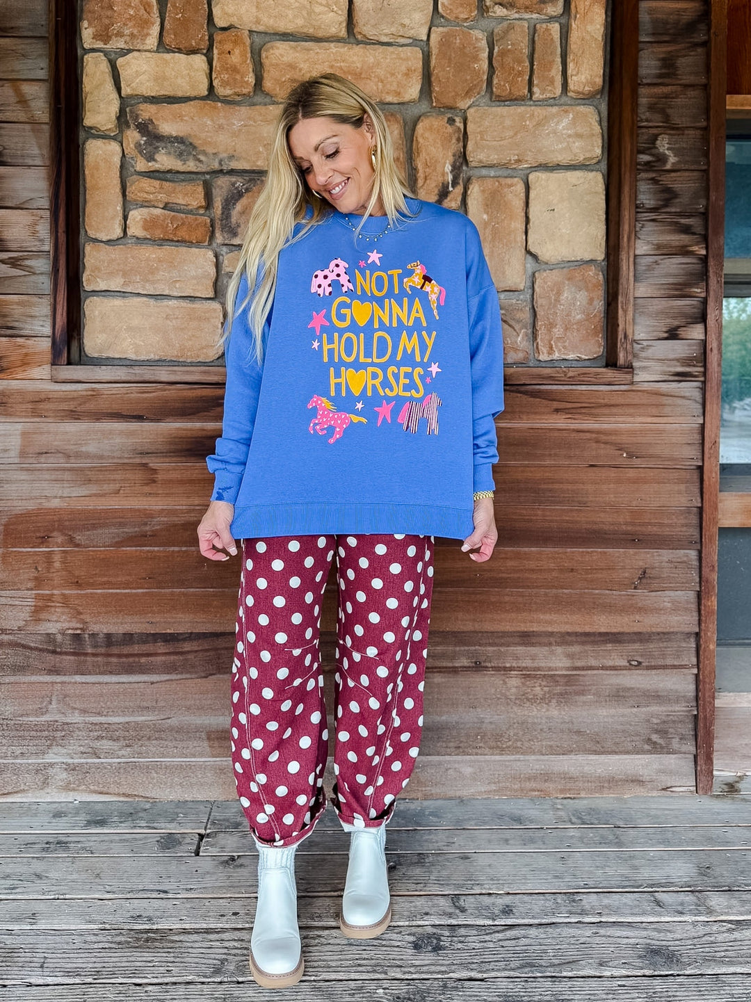 A woman stands on a wooden porch in front of a stone wall, wearing a bright blue sweatshirt with a colorful graphic that reads &