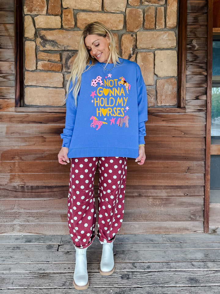 A woman stands on a wooden porch in front of a stone wall, wearing a bright blue sweatshirt with a colorful graphic that reads &