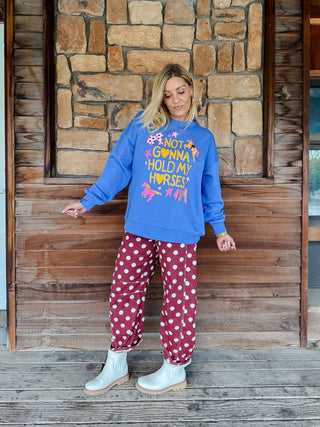 A woman stands on a wooden porch in front of a stone wall, wearing a bright blue sweatshirt with a colorful graphic that reads 'I LOVE YOU HOLD MY HORSY!' and red pants with white polka dots.