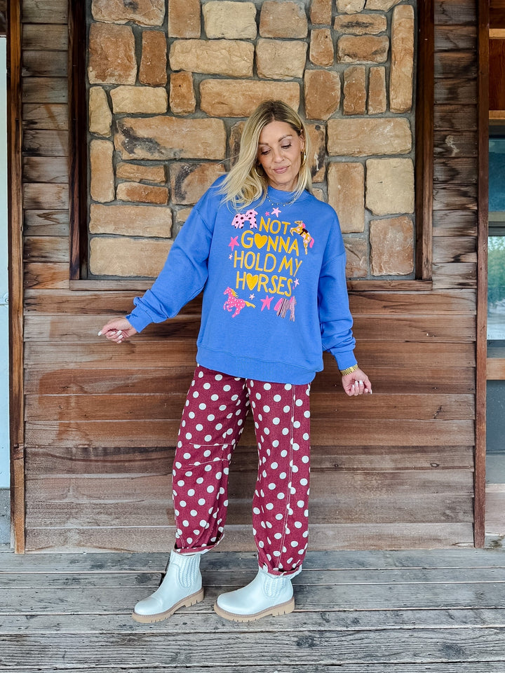 A woman stands on a wooden porch in front of a stone wall, wearing a bright blue sweatshirt with a colorful graphic that reads &