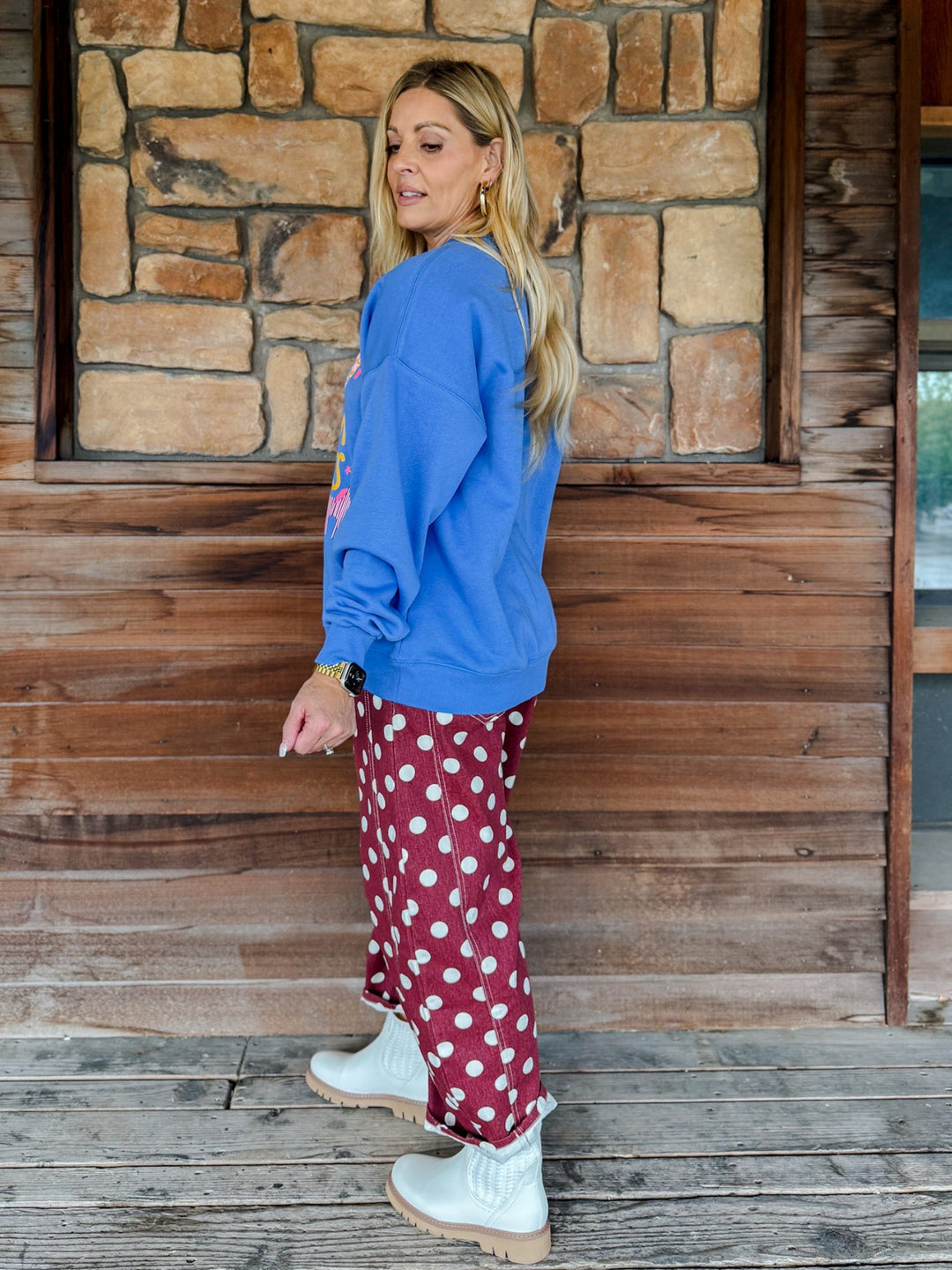 A woman stands on a wooden porch in front of a stone wall, wearing a bright blue sweatshirt, burgundy pants with white polka dots, and white boots.