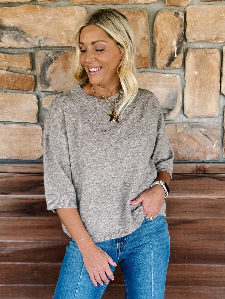 A smiling blonde woman wearing a heather gray knit sweater and blue jeans, standing in front of a rustic stone wall and wooden bench.