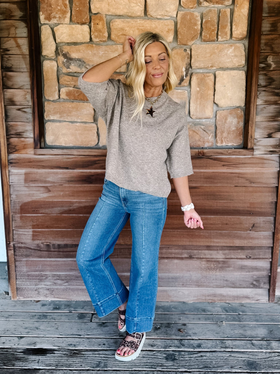 A woman posing on a rustic wooden porch in front of a stone wall, wearing a heathered taupe short-sleeve knit top, blue wide-leg jeans, and patterned sandals.