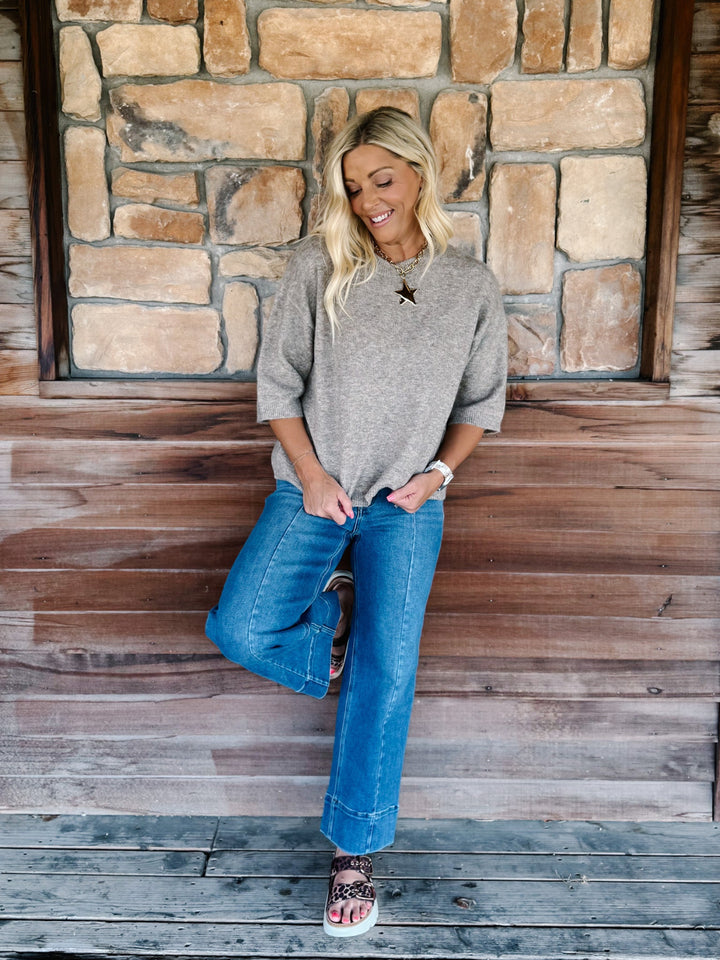 A woman posing on rustic wooden porch steps in front of a stone wall, wearing a heather gray knit sweater, blue jeans, and brown sandals.