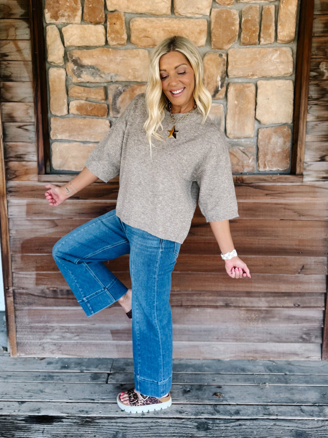 A woman posing in a cozy setting, wearing a heathered taupe knit short-sleeve top, blue jeans, and patterned slip-on shoes, standing in front of a rustic stone and wood wall.