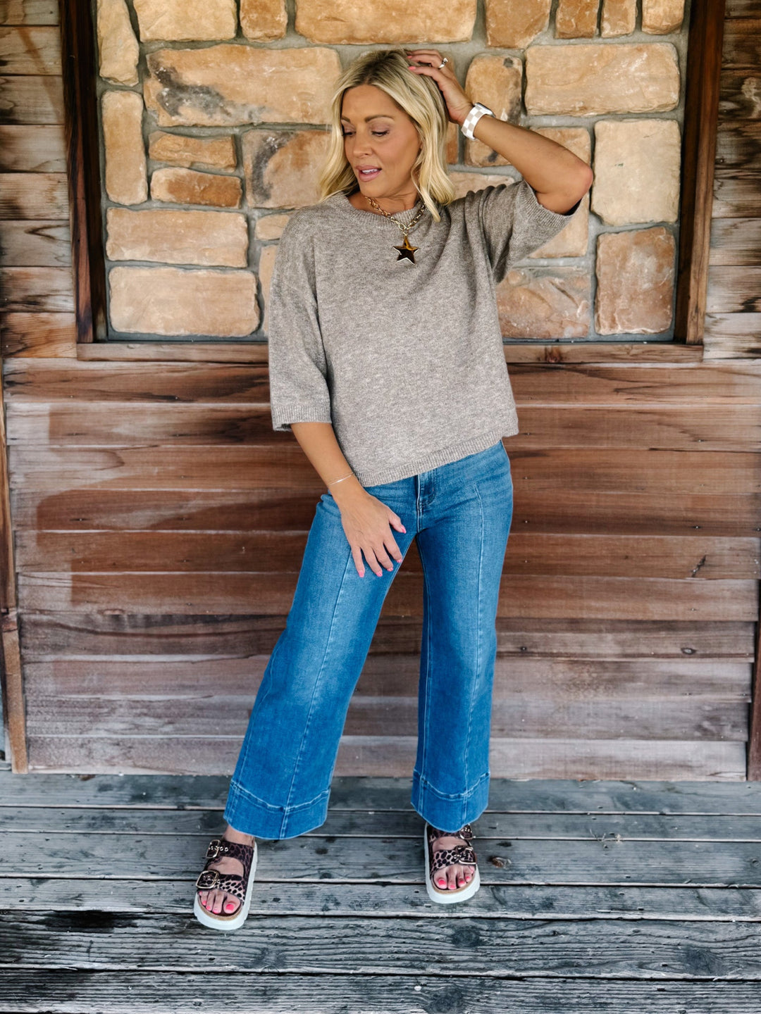 A woman stands on a rustic wooden porch in front of a stone wall, wearing a loose heather gray knit top, blue wide-leg jeans, and pink-and-black patterned sandals.