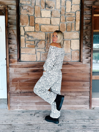 A woman wearing a matching off-the-shoulder long-sleeve top and wide-leg pants set in a black and white abstract pattern, posing on a rustic wooden porch with a stone wall background.