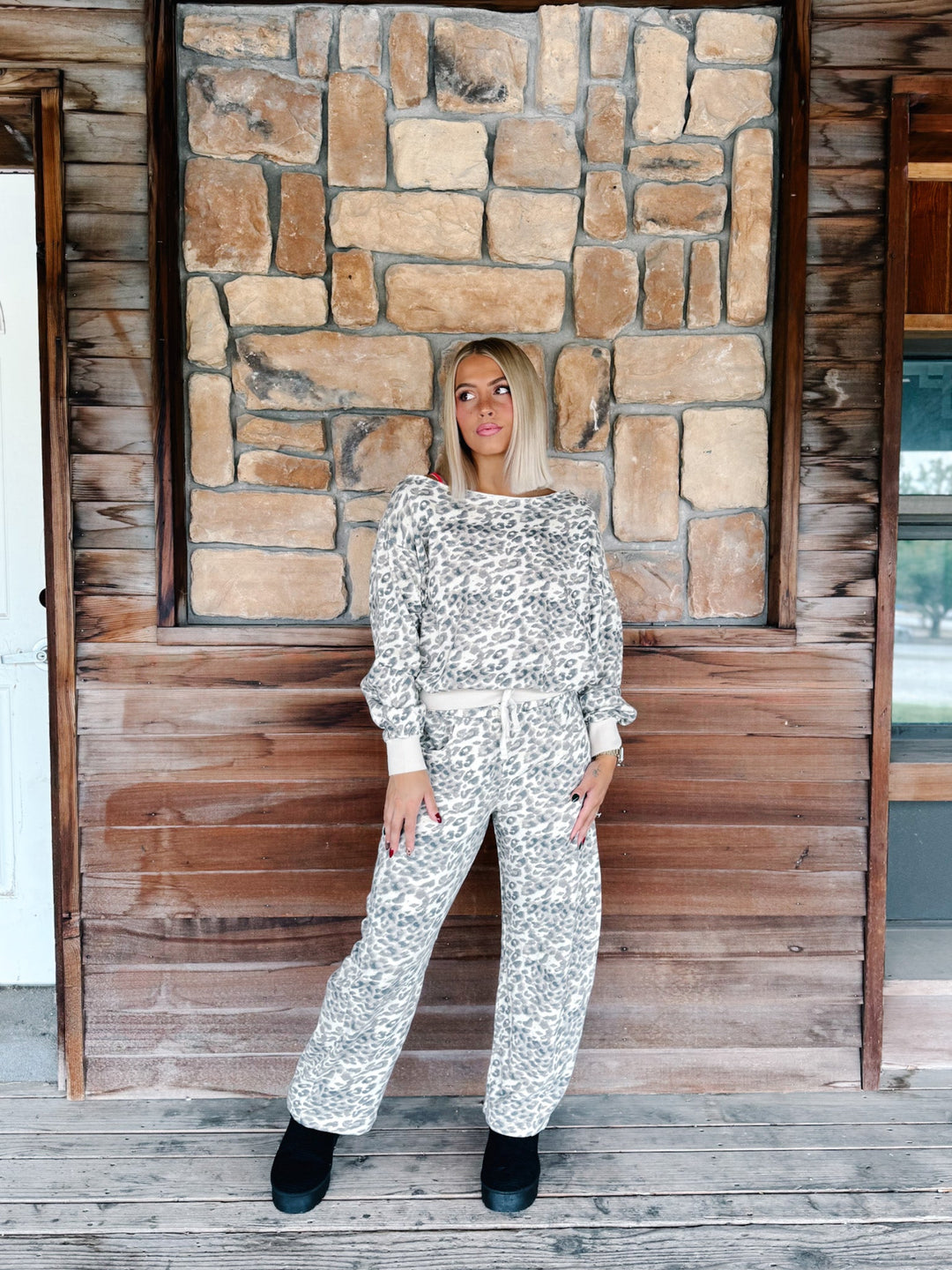 A woman modeling a matching two-piece outfit with a white and gray abstract pattern, standing on a rustic wooden porch in front of a stone wall.