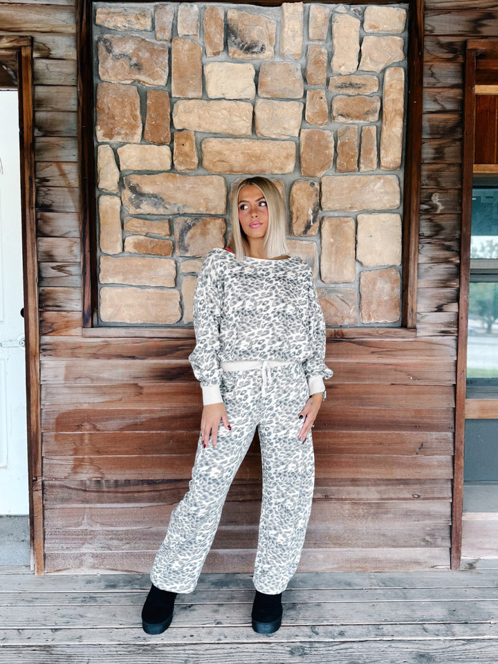 A woman modeling a matching two-piece outfit with a white and gray abstract pattern, standing on a rustic wooden porch in front of a stone wall.