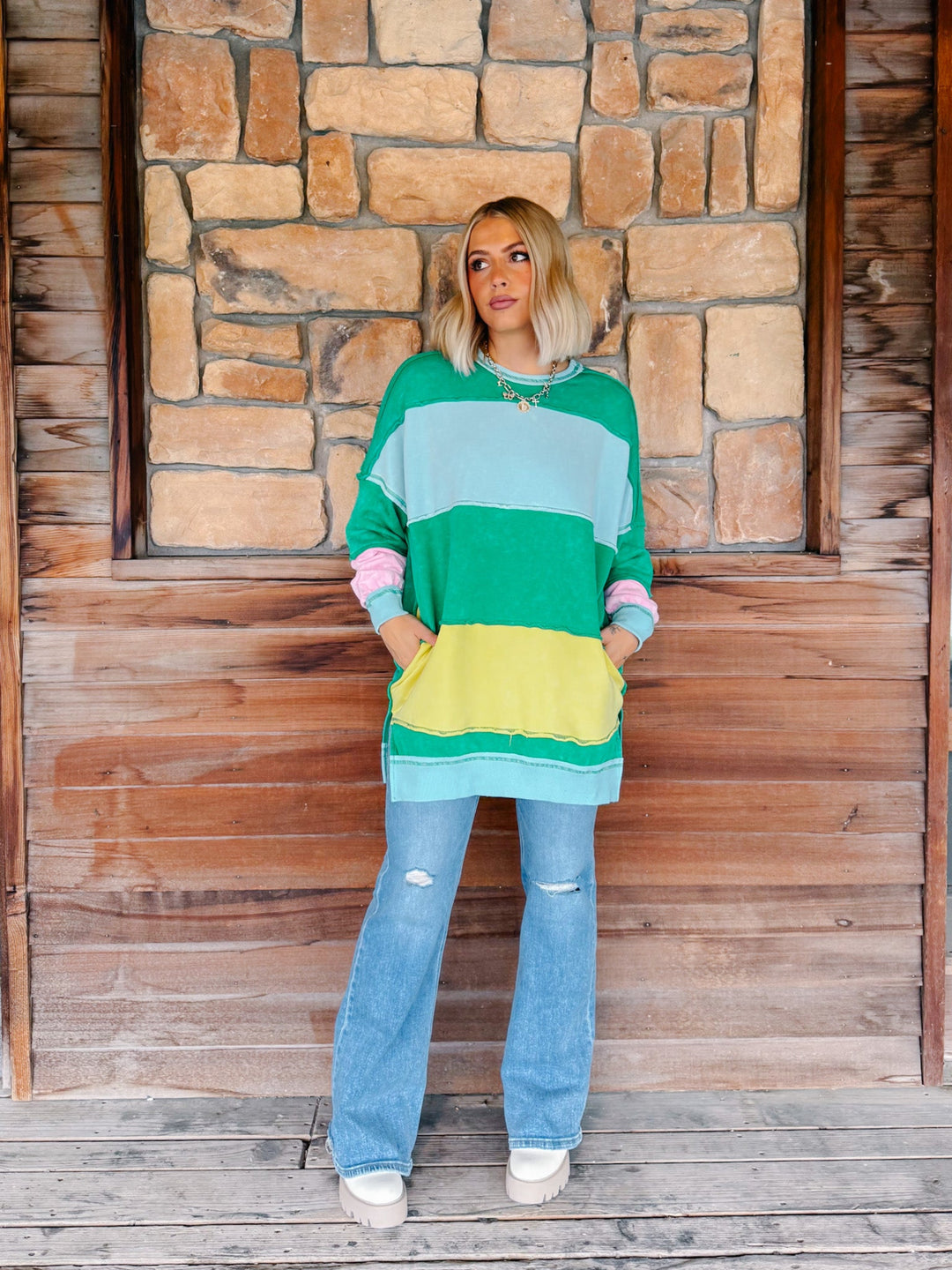 A woman stands on a wooden porch in front of a stone wall, wearing a colorful striped long-sleeve tunic sweater with pockets, light-wash flare jeans, and white sneakers.