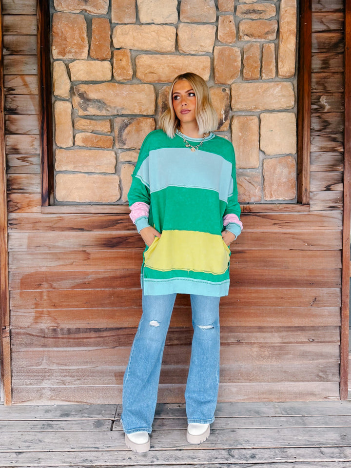 A woman stands on a wooden porch in front of a stone wall, wearing a colorful striped long-sleeve tunic sweater with pockets, light-wash flare jeans, and white sneakers.