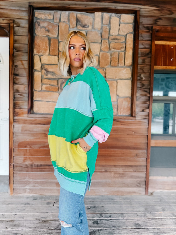 A woman wearing a colorful color-block sweatshirt with green, light blue, yellow, and pink sections, paired with light-wash blue jeans, standing in front of a rustic wooden and stone wall.