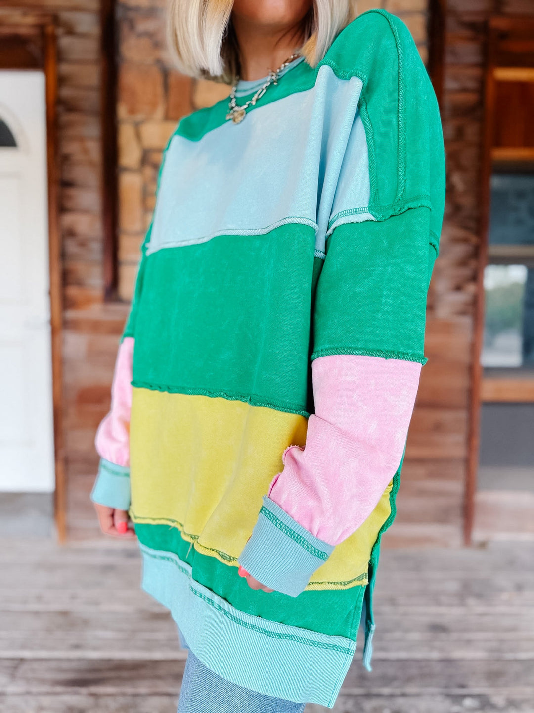 A person wearing a long-sleeve color-block sweatshirt with horizontal stripes of green, light blue, yellow, and pink, photographed in front of a rustic wooden building.