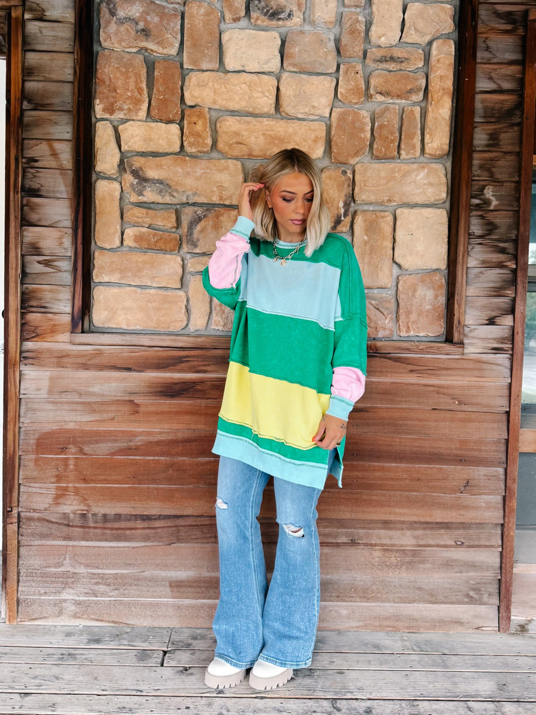 A woman stands on a wooden deck in front of a rustic stone wall, wearing a colorful oversized color-block sweatshirt with green, light blue, yellow, and pink sections, paired with light-wash distressed flare jeans and white sneakers.