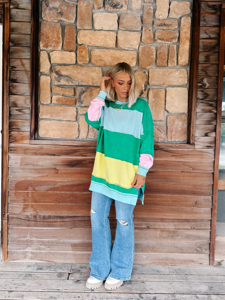 A woman stands on a wooden deck in front of a rustic stone wall, wearing a colorful oversized color-block sweatshirt with green, light blue, yellow, and pink sections, paired with light-wash distressed flare jeans and white sneakers.