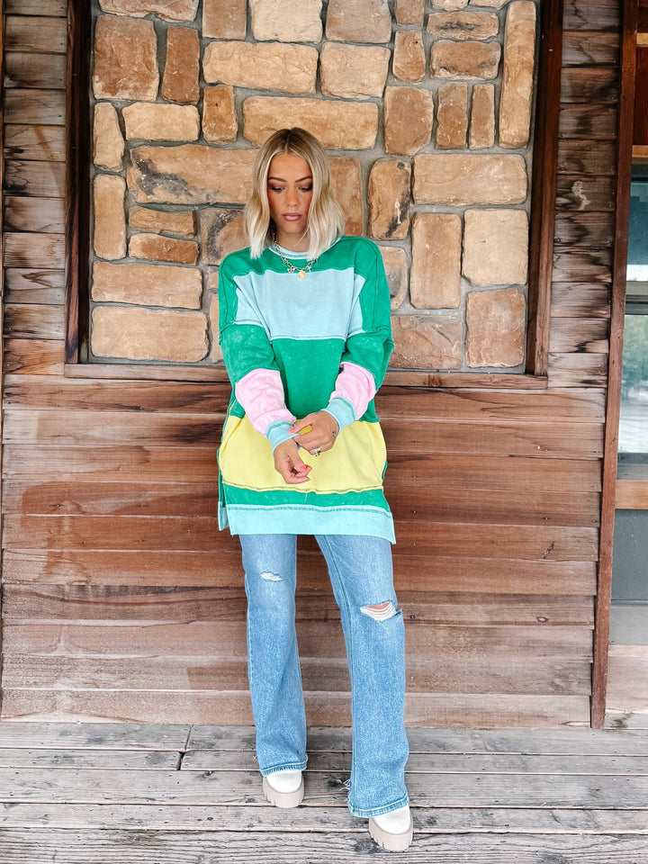 A woman stands on a rustic wooden porch in front of a stone wall, wearing a colorful color-blocked sweatshirt with green, light blue, pink, and yellow panels, paired with light-wash distressed jeans and white sneakers.