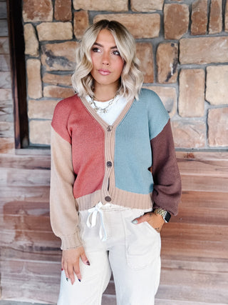 A woman wearing a color-block knit cardigan sweater with beige, dusty pink, light blue, and brown panels, styled with cream drawstring pants against a rustic stone wall background.