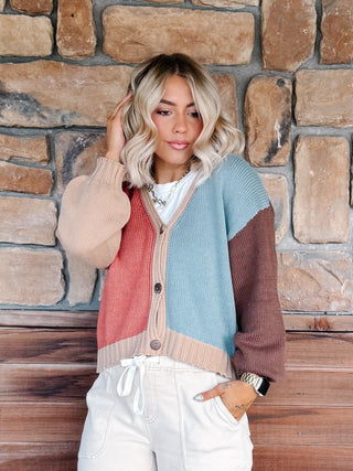 A woman wearing a color-blocked knit cardigan sweater with beige, dusty pink, light blue, and brown sections, paired with white drawstring pants, standing in front of a rustic stone wall.