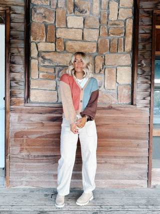 A woman wearing a color-blocked knit cardigan sweater with beige, dusty pink, light blue, and brown sections, paired with white drawstring pants, standing in front of a rustic stone wall.