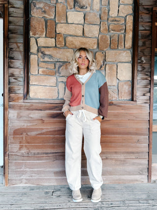 A woman wearing a color-blocked knit cardigan sweater with beige, dusty pink, light blue, and brown sections, paired with white drawstring pants, standing in front of a rustic stone wall.