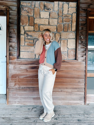 A woman wearing a color-blocked knit cardigan sweater with beige, dusty pink, light blue, and brown sections, paired with white drawstring pants, standing in front of a rustic stone wall.