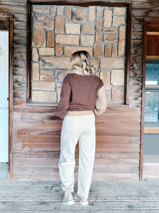 A woman wearing a color-blocked knit cardigan sweater with beige, dusty pink, light blue, and brown sections, paired with white drawstring pants, standing in front of a rustic stone wall.