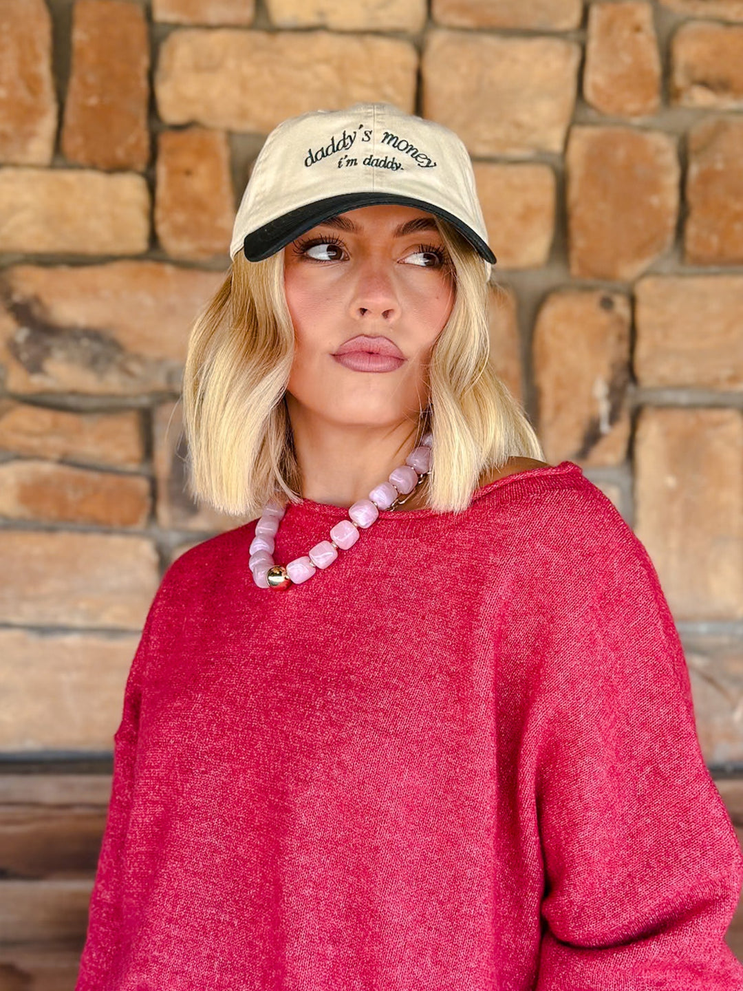 A woman with blonde bob-cut hair wearing a cream baseball cap with black embroidery that reads &