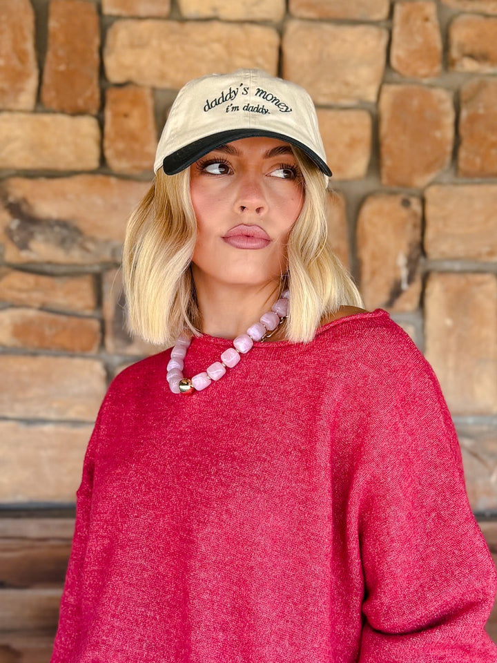 A woman with blonde bob-cut hair wearing a cream baseball cap with black embroidery that reads &