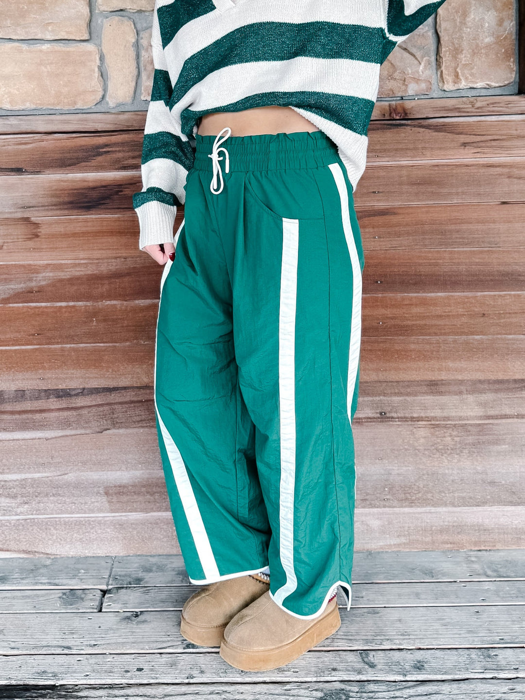 A person wearing green wide-leg pants with white side stripes, paired with a green and white striped sweater and tan slippers, standing against a rustic wooden wall.