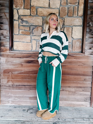 A woman stands on a rustic wooden porch in front of a stone wall, wearing a green and white striped long-sleeve crop top and matching green pants with white side stripes.