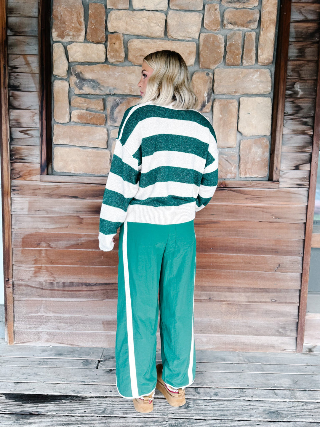 A woman stands with her back to the camera wearing a green and white striped knit sweater and matching green wide-leg track pants with white side stripes, set against a rustic stone and wood wall.