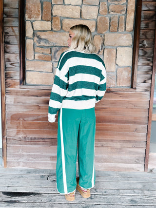 A woman stands with her back to the camera wearing a green and white striped knit sweater and matching green wide-leg track pants with white side stripes, set against a rustic stone and wood wall.