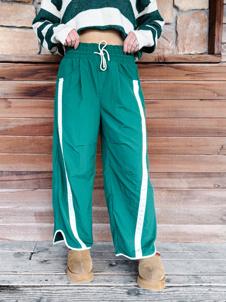 A person wearing green wide-leg athletic pants with white side stripes and a white drawstring, paired with a green-and-white striped sweater and tan boots, standing against a rustic wooden wall.