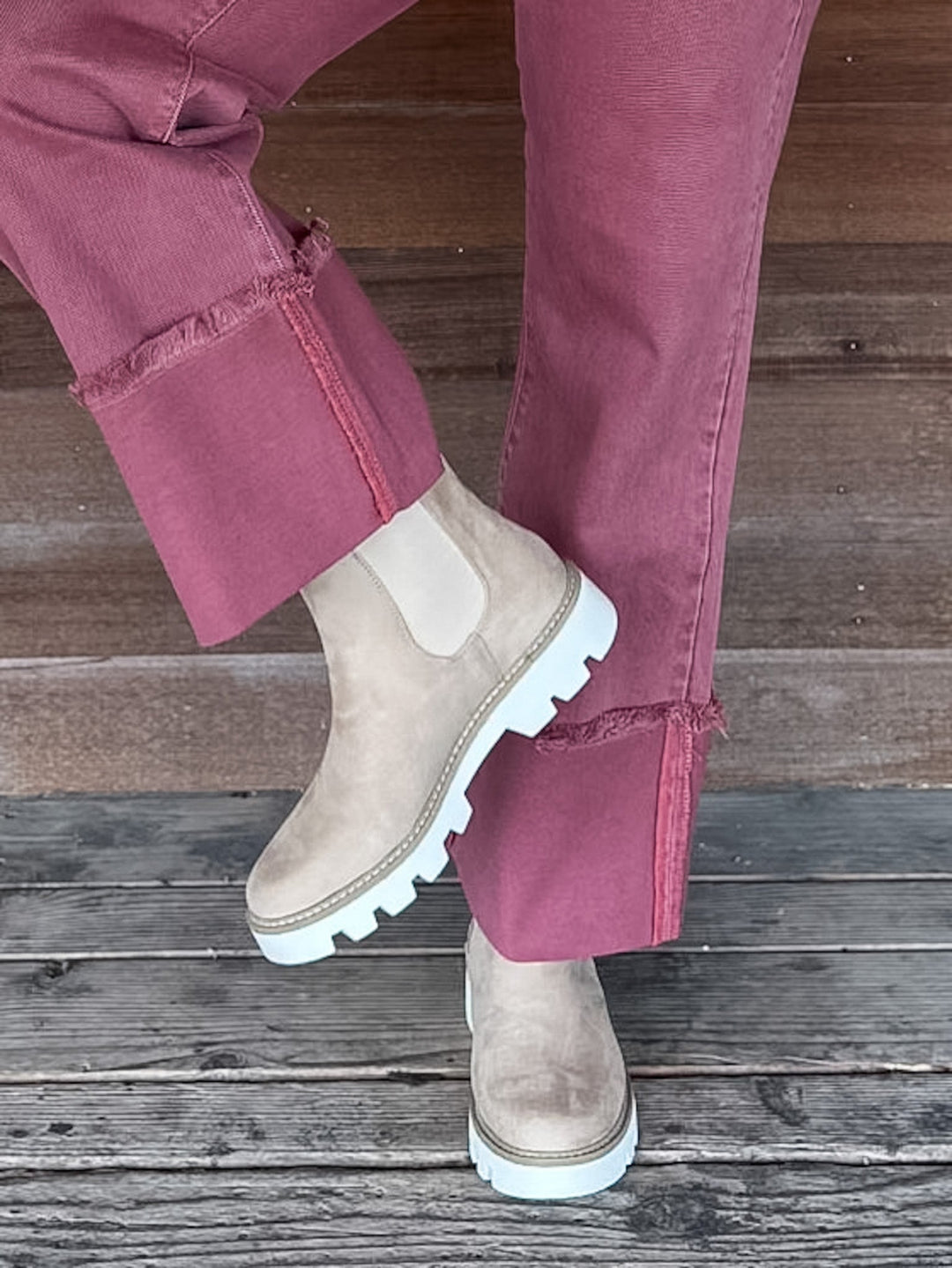 A person wearing dusty rose rolled-up jeans and light beige suede Chelsea-style boots with white soles, standing on weathered wooden planks.
