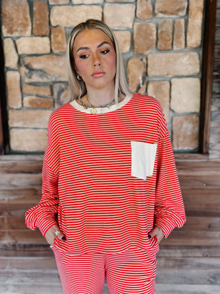 A woman wearing a red and cream striped long-sleeve top with a white chest pocket, paired with matching striped pants, standing in front of a rustic stone wall.