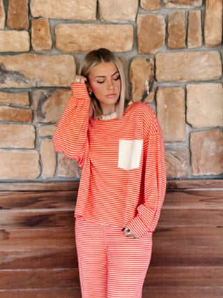 A woman wearing a red and cream striped long-sleeve top with a white chest pocket, paired with matching striped pants, standing in front of a rustic stone wall.