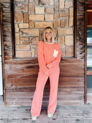 A woman wearing a red and cream striped long-sleeve top with a white chest pocket, paired with matching striped pants, standing in front of a rustic stone wall.