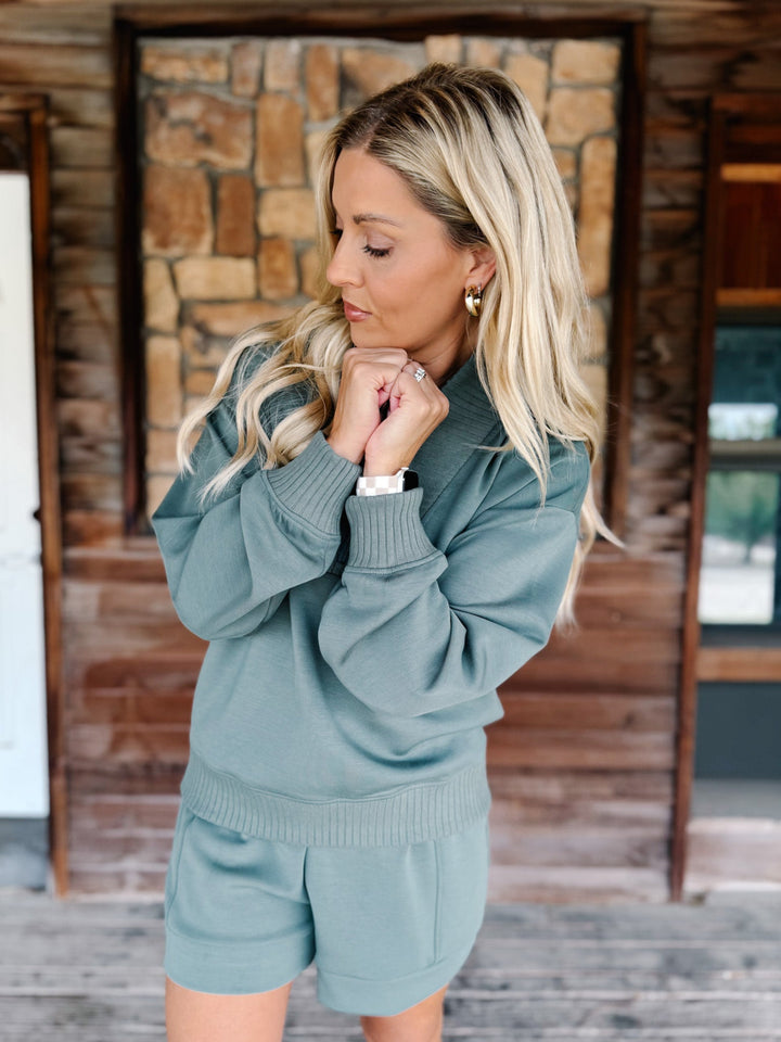 A woman wearing a matching sage green knit lounge set consisting of a long-sleeve collared sweatshirt and shorts, standing on a rustic wooden porch in front of a stone fireplace.