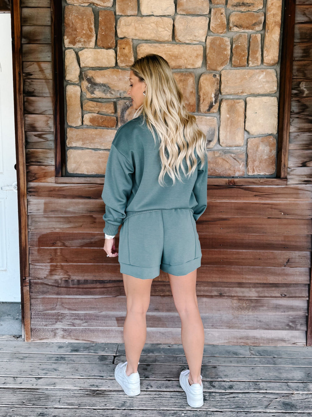 A woman stands on a wooden porch wearing a sage green two-piece lounge set consisting of a long-sleeve sweatshirt and matching shorts, paired with white sneakers.